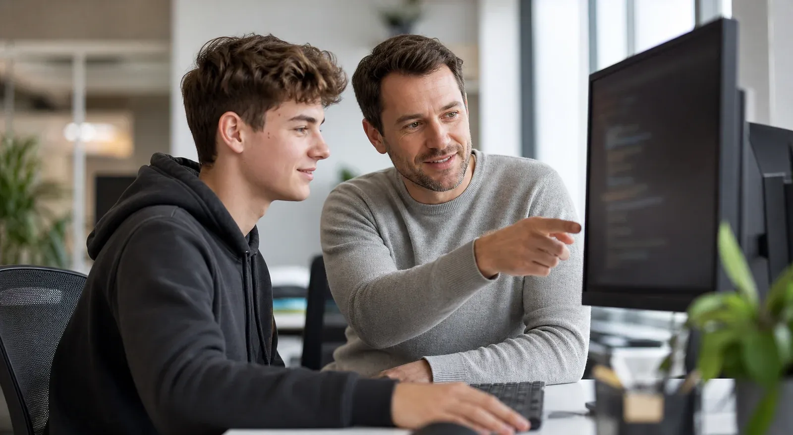 Junger Entwickler und erfahrener Kollege besprechen gemeinsam Code an einem Monitor in einem hellen Büro